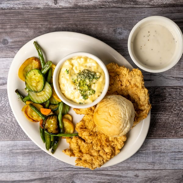 Fried chicken, roasted vegetables, cheesy broccoli, a dinner roll, and gravy on a white plate.