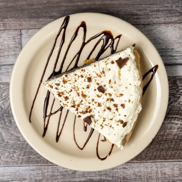 Slice of pie with whipped cream and chocolate shavings, drizzled with chocolate syrup on a plate.